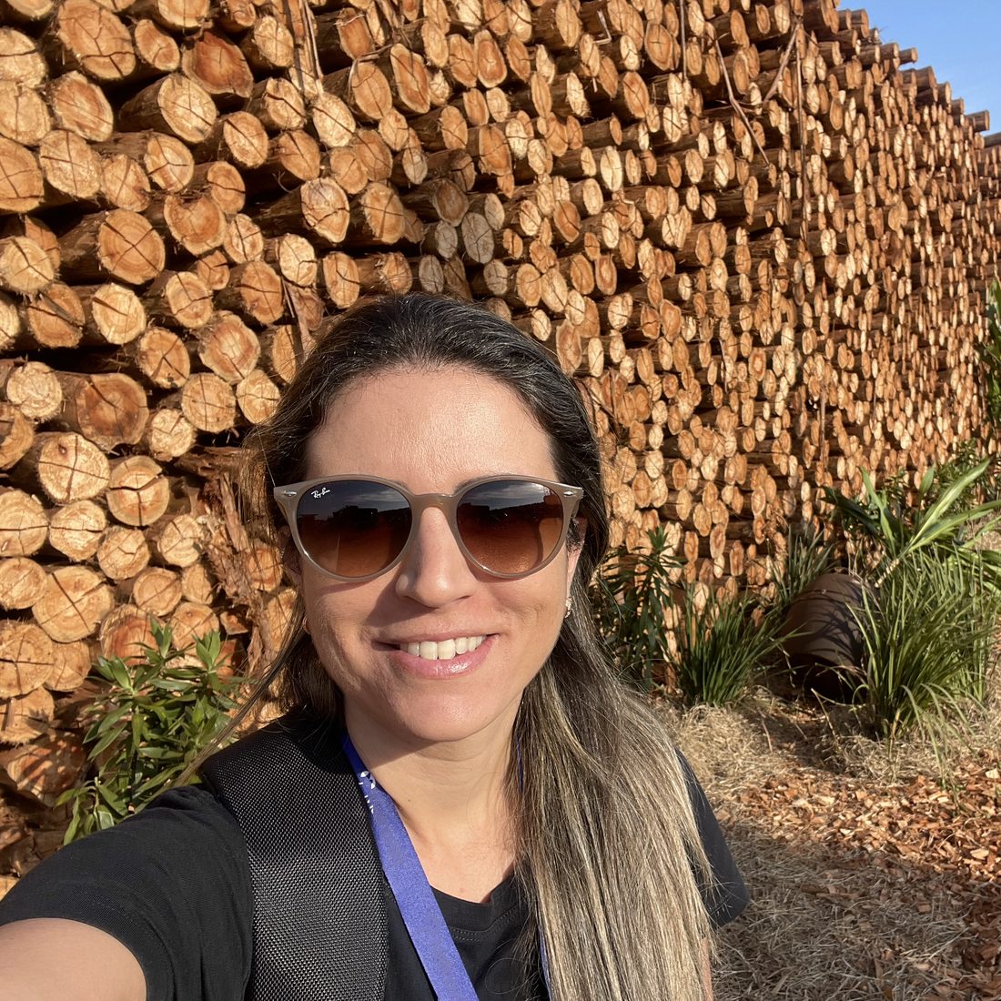 Mila Liparize de Oliveira em pátio de madeira, diante de pilha de toras de eucalipto